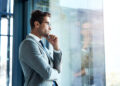 A man in a gray suit and glasses stands by a large window, looking thoughtful with his hand on his chin. His reflection is visible in the glass. | FintechZoom