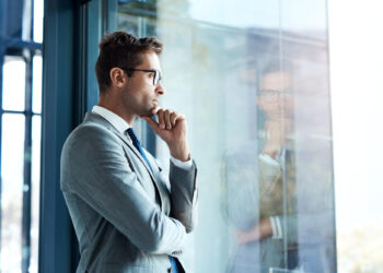 A man in a gray suit and glasses stands by a large window, looking thoughtful with his hand on his chin. His reflection is visible in the glass. | FintechZoom