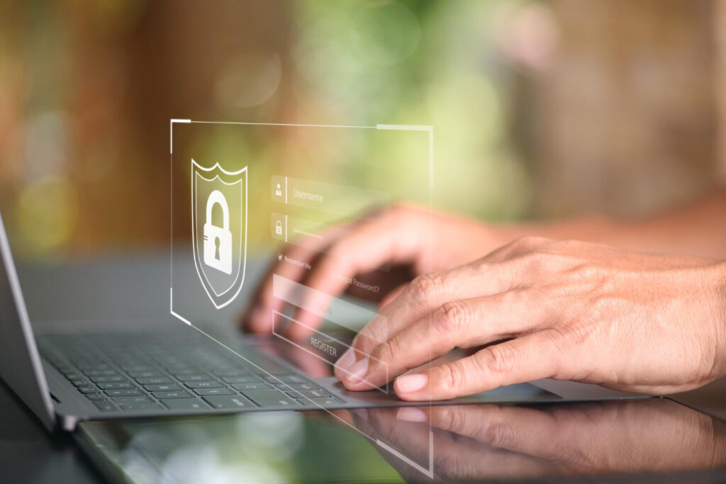 A person types on a laptop keyboard with a digital shield and padlock icon overlay, symbolizing cybersecurity and secure login. The screen also shows blurred login fields for username and password. | FintechZoom