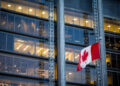A Canadian flag waves in front of a modern glass office building with rows of illuminated windows at dusk. | FintechZoom