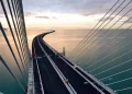 A cable-stayed bridge curves over calm, blue-green water at sunset. The sky is partly cloudy with warm light reflecting on the surface. The bridge is empty, and suspension cables frame the scenic view. | FintechZoom