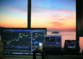Multiple screens display stock market charts and graphs on a desk near a window, with a colorful sunset and calm water visible outside. | FintechZoom