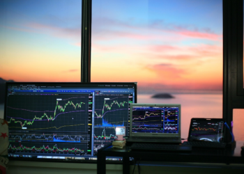 Multiple screens display stock market charts and graphs on a desk near a window, with a colorful sunset and calm water visible outside. | FintechZoom