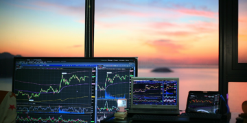 Multiple screens display stock market charts and graphs on a desk near a window, with a colorful sunset and calm water visible outside. | FintechZoom