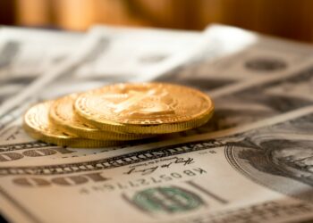 Three gold Bitcoin coins are stacked on top of several one hundred dollar US bills, highlighting the contrast between cryptocurrency and traditional currency. The background is softly blurred. | FintechZoom