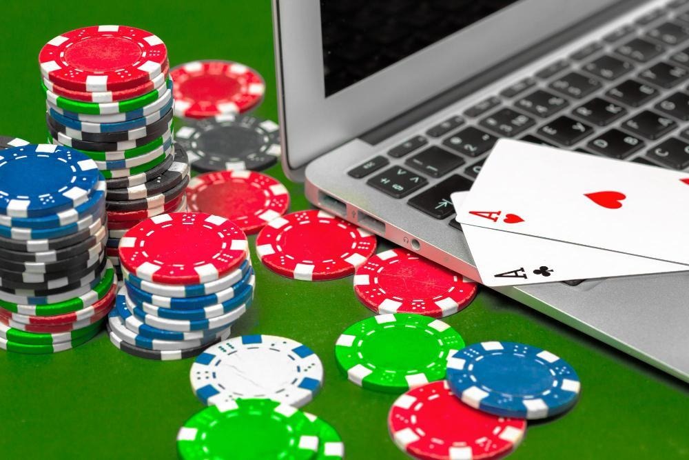 Poker chips and two playing cards (ace of hearts and ace of clubs) are next to an open laptop on a green table, representing online gambling or poker. | FintechZoom