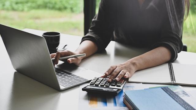 A person working at a desk using a laptop and a calculator, with a notebook, pen, documents, and a cup nearby. The scene appears to be in a bright, modern workspace with a window in the background. | FintechZoom