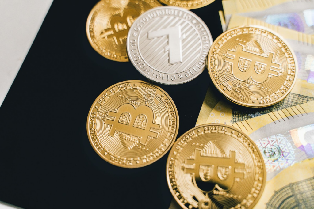 Several gold Bitcoin coins and one silver Litecoin coin are arranged on a black surface next to euro banknotes. | FintechZoom