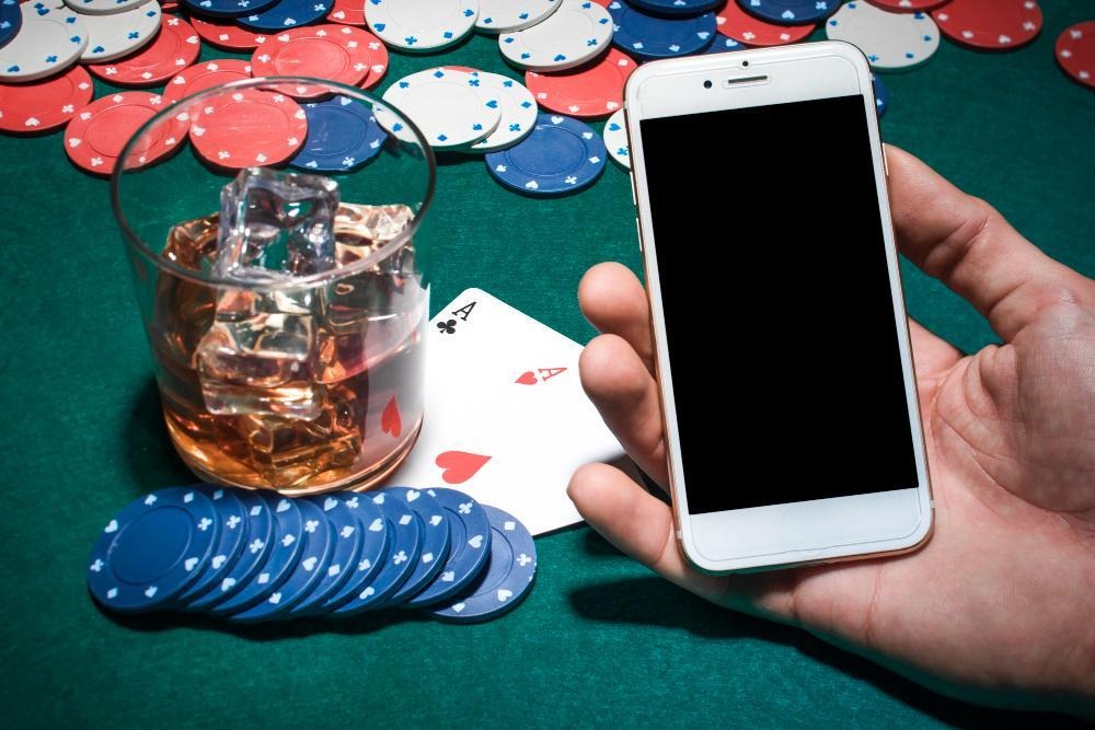 A hand holds a smartphone with a blank screen next to a glass of whiskey with ice, poker chips, and two playing cards (ace of hearts and ace of clubs) on a green poker table. | FintechZoom