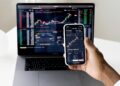 A person holds a smartphone displaying a stock trading app with charts, in front of a laptop showing similar financial graphs and data, indicating active stock market monitoring. | FintechZoom