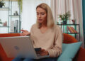 A woman with blonde hair sits on an orange couch, holding a credit card and gesturing at her laptop screen. She appears to be making an online purchase or attending a video call in a home setting with plants and shelves in the background. | FintechZoom
