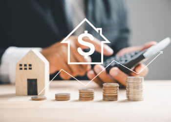 A person in a suit uses a calculator next to a small wooden house and stacks of coins. A dollar sign inside a house icon and a rising graph are overlaid, symbolizing real estate investment and financial growth. | FintechZoom
