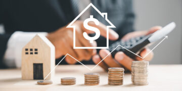 A person in a suit uses a calculator next to a small wooden house and stacks of coins. A dollar sign inside a house icon and a rising graph are overlaid, symbolizing real estate investment and financial growth. | FintechZoom