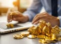 A person in business attire uses a laptop with stacks of gold coins and gold nuggets on the desk, suggesting finance, investment, or trading activities. | FintechZoom