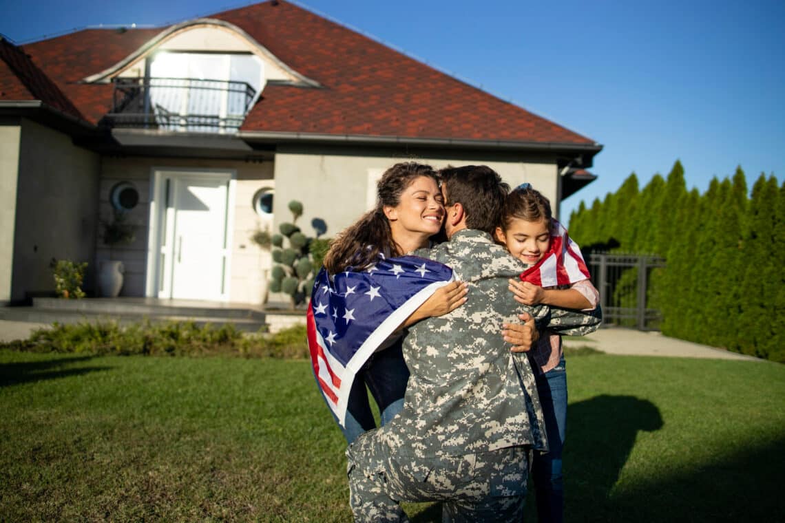 A soldier in uniform hugs his smiling wife and young daughter outside their house. Wrapped in American flags, the family looks thrilled to be reunited—thanks to the Best VA Mortgage options that helped them find their perfect home. | FintechZoom