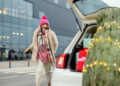 A woman in a pink hat, colorful scarf, and fur coat walks happily toward a white car with its trunk open, revealing a decorated Christmas tree and wrapped gifts inside, outside a modern building. | FintechZoom