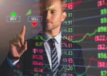A businessman in a suit touches a transparent virtual screen with "BUY" and "SELL" buttons, while colorful stock market graphs and numbers are displayed in the background. | FintechZoom