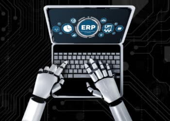 A robot with metallic hands types on a laptop keyboard displaying an ERP (Enterprise Resource Planning) interface and various business icons, set against a dark, tech-themed background. | FintechZoom