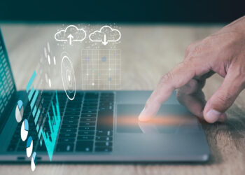 A hand uses the trackpad on a laptop, with digital graphics of cloud computing, data charts, and futuristic interface elements overlayed above the keyboard. | FintechZoom