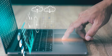 A hand uses the trackpad on a laptop, with digital graphics of cloud computing, data charts, and futuristic interface elements overlayed above the keyboard. | FintechZoom