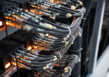 Close-up of several bundles of network cables connected to a server rack, with orange indicator lights glowing, illustrating data transmission or network infrastructure in a server room. | FintechZoom