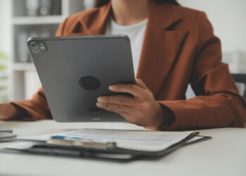A person in a brown blazer sits at a desk, holding a tablet. Papers, a clipboard, and a pen are on the desk in front of them. The scene appears to be in an office or professional setting. | FintechZoom
