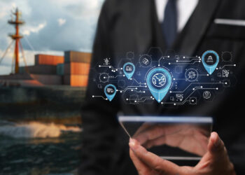 A person in a suit holds a tablet with digital icons overlay, representing logistics and global shipping, in front of a cargo ship loaded with containers at sea. | FintechZoom