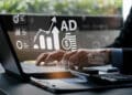 A person types on a laptop with virtual graphics showing charts, a rising arrow, coins, “AD” text, and marketing icons, suggesting digital advertising or online marketing analytics. | FintechZoom