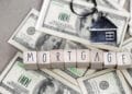Wooden letter blocks spelling "MORTGAGE" rest on top of scattered U.S. hundred-dollar bills, with a metal house-shaped keychain visible in the background. | FintechZoom