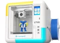 A compact X-Maker 3D printer with a transparent front door shows a blue and yellow robot figure being printed. A spool of blue filament is mounted on the right side of the white and turquoise printer. | FintechZoom