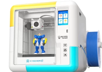 A compact X-Maker 3D printer with a transparent front door shows a blue and yellow robot figure being printed. A spool of blue filament is mounted on the right side of the white and turquoise printer. | FintechZoom