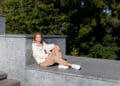 A woman in beige pants, a white jacket, and white sneakers sits on a gray stone platform outdoors with her eyes closed, enjoying the sunlight—a perfect moment for any Sneaker Style Guide. Green trees fill the background. | FintechZoom