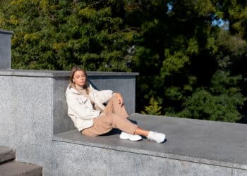 A woman in beige pants, a white jacket, and white sneakers sits on a gray stone platform outdoors with her eyes closed, enjoying the sunlight—a perfect moment for any Sneaker Style Guide. Green trees fill the background. | FintechZoom