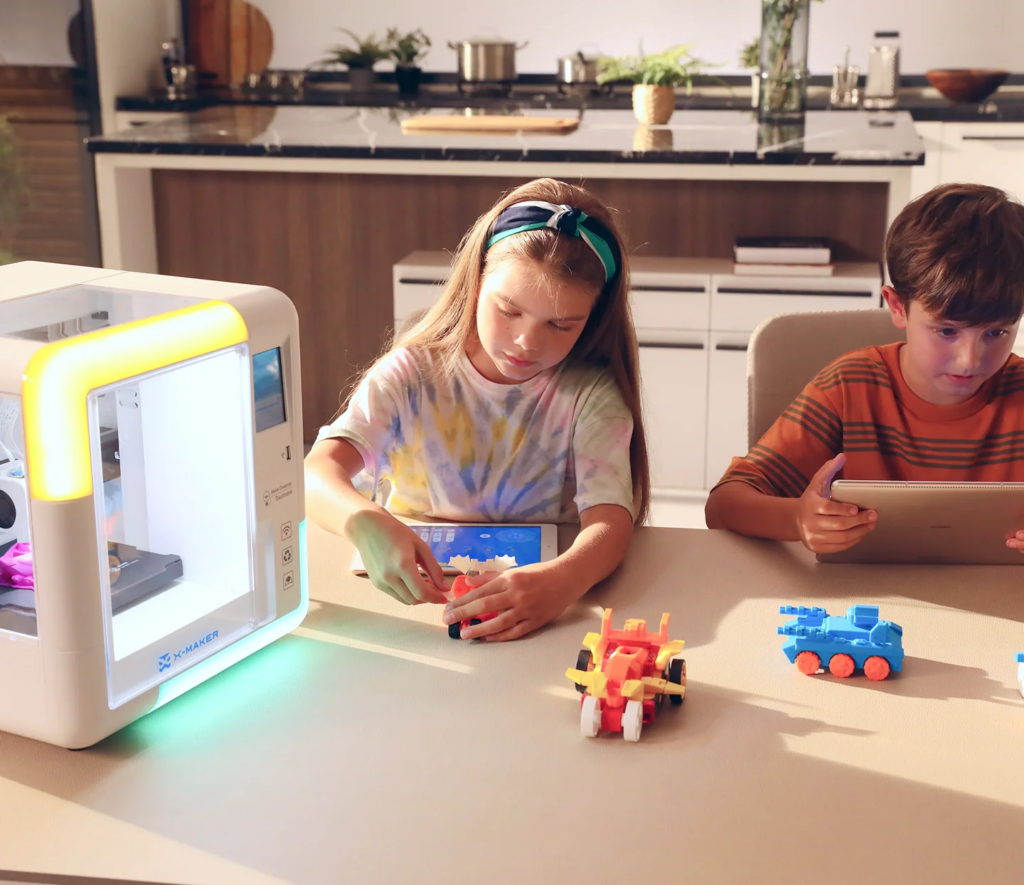 Two children sit at a table in a kitchen, playing with colorful toy vehicles. A 3D printer is on the table, and the girl assembles a toy while the boy uses a tablet. The kitchen is bright and modern. | FintechZoom
