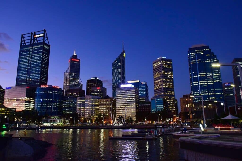  Photo of the waterfront in Perth, Australia : A skyline at dusk with tall, illuminated office buildings reflecting on the water, boats docked in the harbor, and a clear, deep blue sky overhead. | FintechZoom