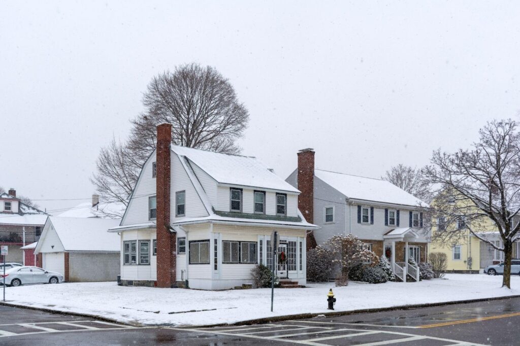 Two houses on a street corner, covered in a light layer of snow, stand quietly as snowflakes gently fall. Bare trees and a fire hydrant line the curb, creating a peaceful winter scene—perfect for those considering a refinance this season. | FintechZoom
