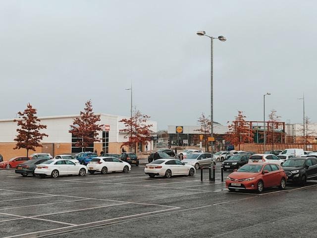 A mostly empty parking lot with several parked cars, bordered by small trees with autumn leaves. In the background, there are commercial buildings, including a Lidl supermarket. The sky is overcast. | FintechZoom