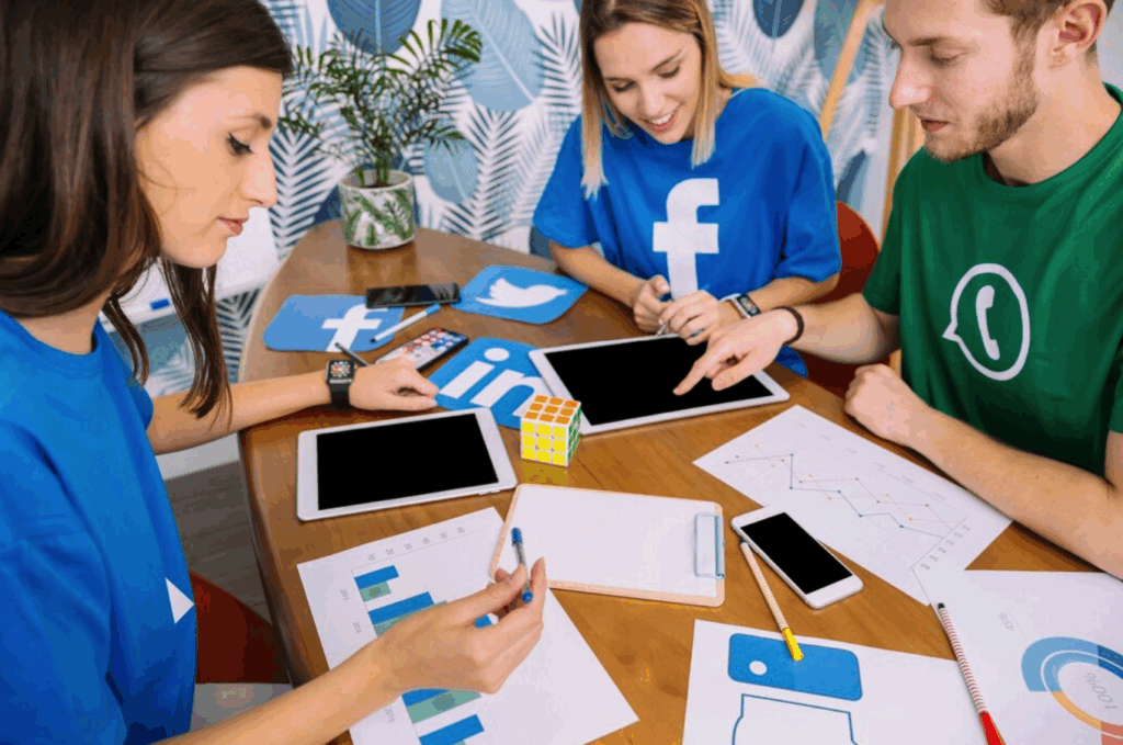 Three young people sit at a table with charts, tablets, and social media Meta-owned companies icons, collaborating on a project. They wear shirts featuring Facebook, WhatsApp, and other social media logos. A Rubik's Cube is on the table. | FintechZoom