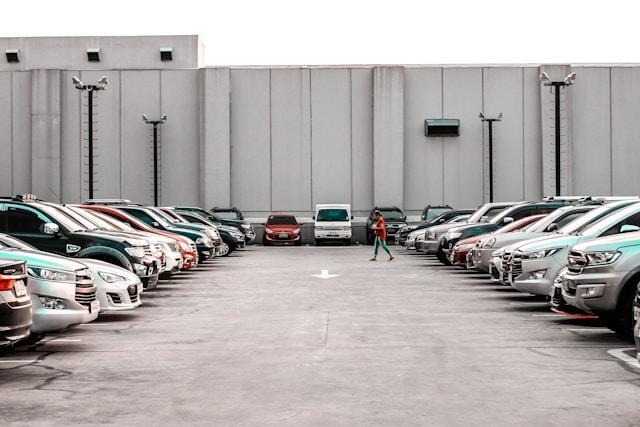 A person walks through a parking lot filled with cars parked in rows on both sides, with a white wall and several light poles in the background. | FintechZoom