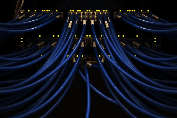 A network server rack with multiple blue Ethernet cables connected to ports. Small yellow indicator lights are lit above each port, highlighting active connections in a data center or networking environment. | FintechZoom