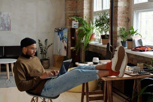 A man wearing a brown sweater, beanie, and glasses sits with his legs up on a desk, working on a laptop in a cozy room filled with plants and natural light. | FintechZoom