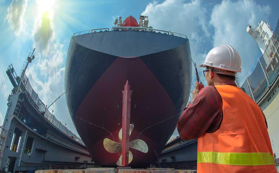 A worker in a white hard hat and orange safety vest inspects the bow of a large ship in dry dock under a partly cloudy sky with sunlight streaming in. | FintechZoom
