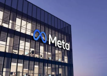 A glass office building at dusk displays the illuminated Meta logo and name on its facade. The interior shows multiple floors with lit offices and furniture visible through the windows. | FintechZoom