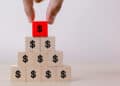 A hand places a red wooden block with a dollar sign on top of a pyramid made of beige blocks, each also marked with a dollar sign, symbolizing financial growth or investment. | FintechZoom