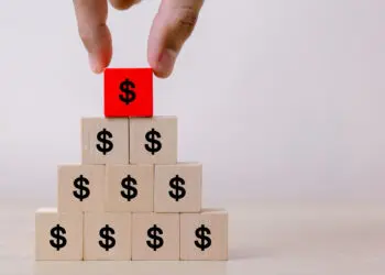 A hand places a red wooden block with a dollar sign on top of a pyramid made of beige blocks, each also marked with a dollar sign, symbolizing financial growth or investment. | FintechZoom