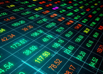 A digital display of stock market prices showing rows of brightly colored numbers in red, green, yellow, and blue on a dark grid background, representing fluctuating financial data. | FintechZoom