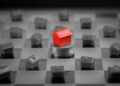 A red toy house stands on stacked coins at the center of a chessboard-like grid, surrounded by small gray houses, symbolizing real estate investment or property value. | FintechZoom