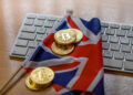 A small Union Jack flag and several gold Bitcoin coins are placed on a computer keyboard atop a wooden desk. | FintechZoom