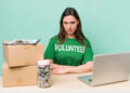 A woman in a green "VOLUNTEER" shirt sits at a desk with a laptop, a jar of cash, two cardboard boxes, and a clipboard, looking seriously at the camera against a light green background. | FintechZoom