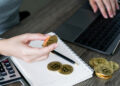A person holds a physical Bitcoin coin near a notebook and pen, with several more Bitcoin coins on the table, while using a laptop and calculator in the background. | FintechZoom